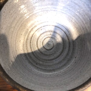 Puede incluir: Un cuenco de cer&aacute;mica hecho a mano con un interior gris claro moteado. El interior del cuenco presenta crestas circulares conc&eacute;ntricas, creando un patr&oacute;n en espiral. El borde es de color marr&oacute;n oscuro. El cuenco se utiliza probablemente para servir alimentos o como pieza decorativa.