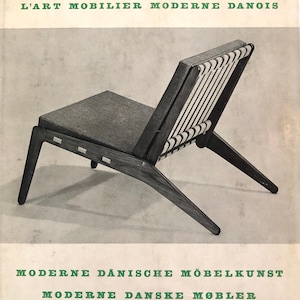 Puede incluir: Una fotograf&iacute;a en blanco y negro de una silla de madera con respaldo tejido y asiento de tela gris. La silla est&aacute; inclinada hacia la derecha y el respaldo de la silla est&aacute; orientado hacia el espectador. La silla est&aacute; sobre un fondo blanco. El texto "MODERN DANISH FURNITURE" est&aacute; en la parte superior de la imagen. El texto "L'ART MOBILIER MODERNE DANOIS" est&aacute; debajo de la primera l&iacute;nea de texto. El texto "MODERNE D&Auml;NISCHE M&Ouml;BELKUNST" est&aacute; en la parte inferior de la imagen. El texto "MODERNE DANSKE M&Oslash;BLER" est&aacute; debajo de la l&iacute;nea de texto anterior. El texto "GJELLERUP" est&aacute; debajo de la l&iacute;nea de texto anterior. El texto "ARCHITECTURAL BOOK PUBLISHING CO. INC. NEW YORK" est&aacute; debajo de la l&iacute;nea de texto anterior.