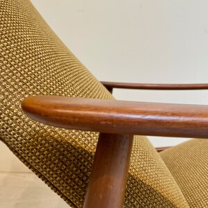 May include: Close-up of a mid-century modern chair. The chair features a wooden frame with a curved armrest and a woven fabric seat and backrest. The fabric is a textured pattern in shades of tan and brown.