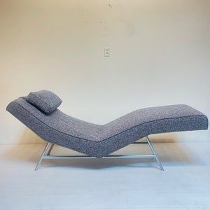 May include: A modern chaise lounge with a gray tweed fabric and a small headrest. The lounge has a curved design and is supported by silver metal legs. The overall design is minimalist and stylish, suitable for a living room or bedroom.