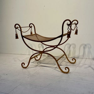 Puede incluir: Un banco de metal dorado con asiento tejido. El banco tiene patas curvas y un respaldo decorativo con borlas. El dise&ntilde;o es ornamentado y de inspiraci&oacute;n vintage, adecuado para uso en interiores.