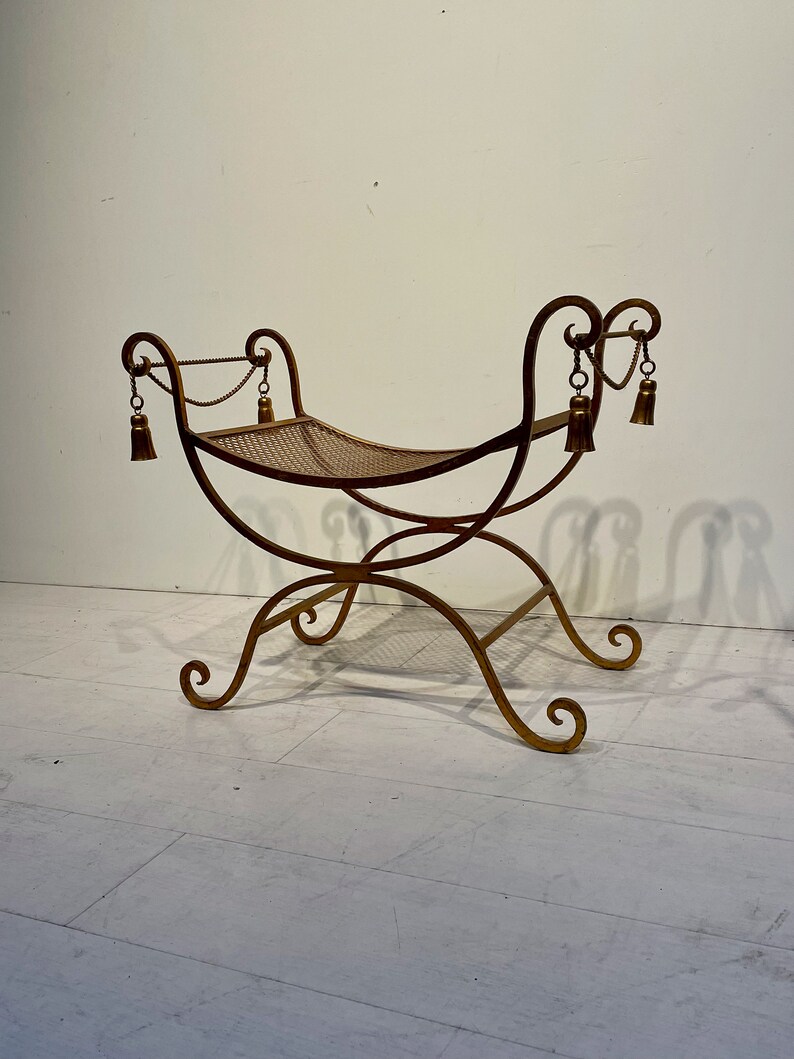 Puede incluir: Un banco de metal dorado con asiento tejido. El banco tiene patas curvas y un respaldo decorativo con borlas. El dise&ntilde;o es ornamentado y de inspiraci&oacute;n vintage, adecuado para uso en interiores.