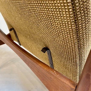 May include: Close-up of a mid-century modern chair. The chair features a wooden frame with a warm brown finish and a woven fabric seat and backrest in shades of olive green and brown. The chair has a minimalist design.