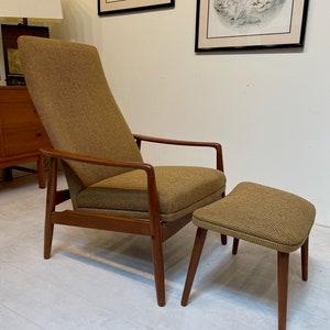 May include: Mid-century modern armchair and matching ottoman. The chair has a high back and wooden armrests. Both pieces are upholstered in a textured, olive-green fabric. The wooden frames have a warm, brown finish. The ottoman is a low, rectangular shape.