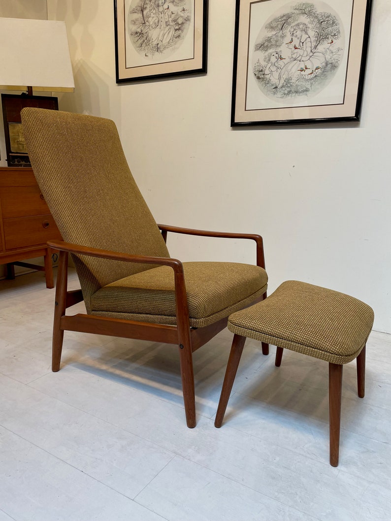 May include: Mid-century modern armchair and matching ottoman. The chair has a high back and wooden armrests. Both pieces are upholstered in a textured, olive-green fabric. The wooden frames have a warm, brown finish. The ottoman is a low, rectangular shape.