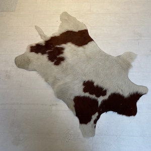 May include: A cowhide rug with a natural shape. The rug features a white background with irregular brown patches. The hide is laid on a light-colored wooden floor, showcasing its texture and pattern. The rug is a decorative floor covering.