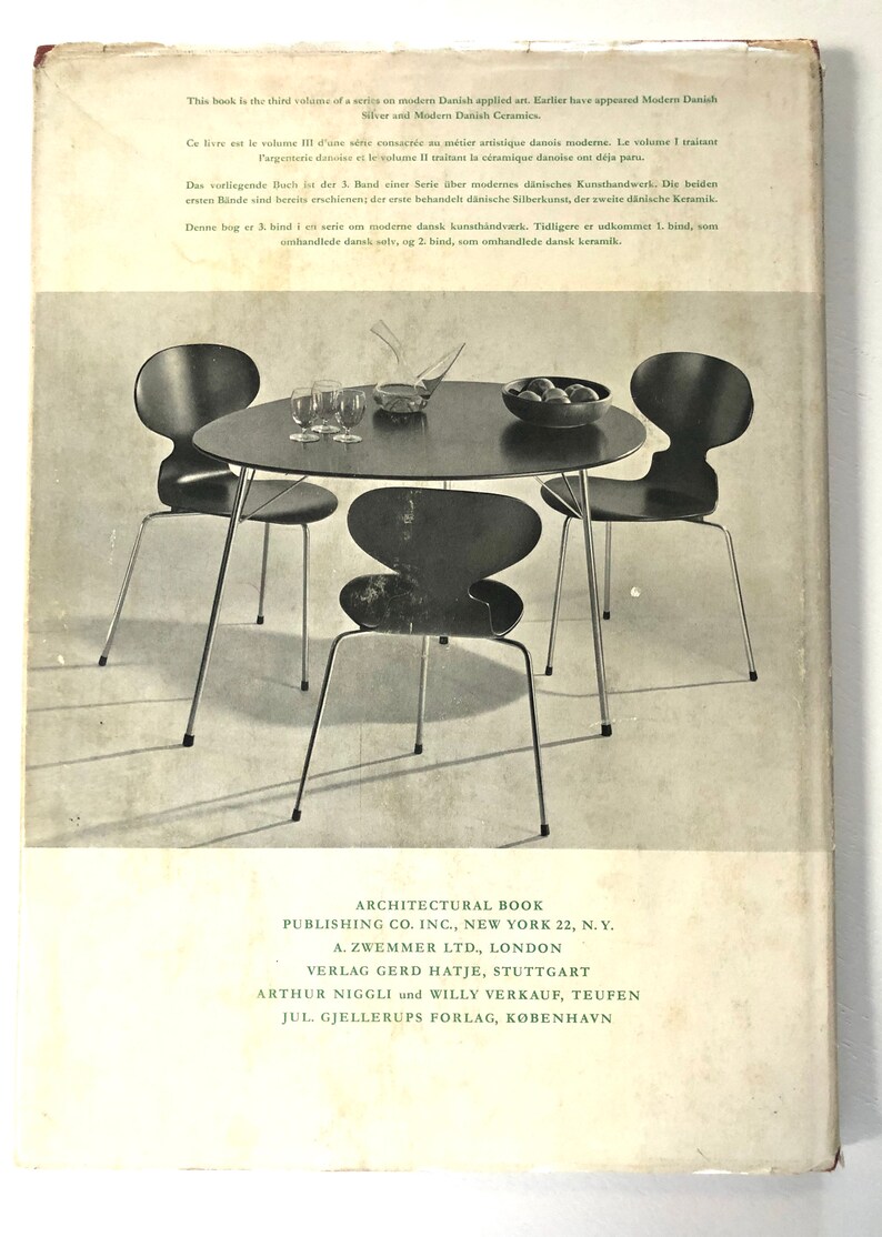 Puede incluir: Una fotograf&iacute;a en blanco y negro de una mesa redonda con tres sillas. La mesa es de madera y tiene una tapa negra. Las sillas son de metal y tienen asientos negros. Las sillas est&aacute;n dispuestas alrededor de la mesa. La mesa y las sillas est&aacute;n en una habitaci&oacute;n con un suelo de color claro. El texto en la parte posterior del libro dice: "Architectural Book Publishing Co. Inc., New York 22, N.Y. A. Zwemmer Ltd., London Verlag Gerd Hatje, Stuttgart Arthur Niggli und Willy Verkauf, Teufen Jul. Gjellerups Forlag, K&oslash;benhavn"