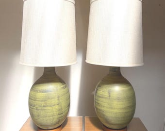 Puede incluir: Un par de lámparas de mesa con bases de cerámica redondeadas en color verde oliva y un acabado texturizado de pinceladas horizontales. Cada lámpara se asienta sobre una base circular de madera y tiene una pantalla de tela cónica de color blanco roto.