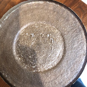 Puede incluir: Primer plano de la base de un jarr&oacute;n de cer&aacute;mica marr&oacute;n. La base circular tiene una superficie texturizada y el texto "The Kin" est&aacute; estampado en el centro.