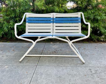 May include: A white metal bench with blue and white horizontal slats. The bench has curved armrests and a simple, modern design. The bench is outdoors, set against a backdrop of green foliage.
