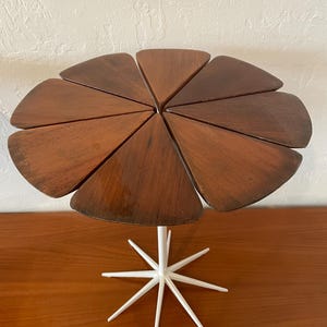 May include: A unique, mid-century modern side table. The tabletop is composed of triangular wooden segments arranged in a flower-like pattern. It is supported by a white, starburst-shaped base. The table is approximately 24 inches in diameter.