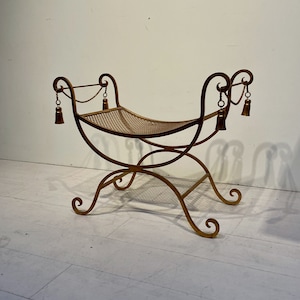 Puede incluir: Un banco de metal dorado con asiento tejido. El banco tiene patas curvas y un respaldo decorativo con borlas colgando de cadenas. El dise&ntilde;o es ornamentado y elegante, adecuado para la decoraci&oacute;n de interiores.