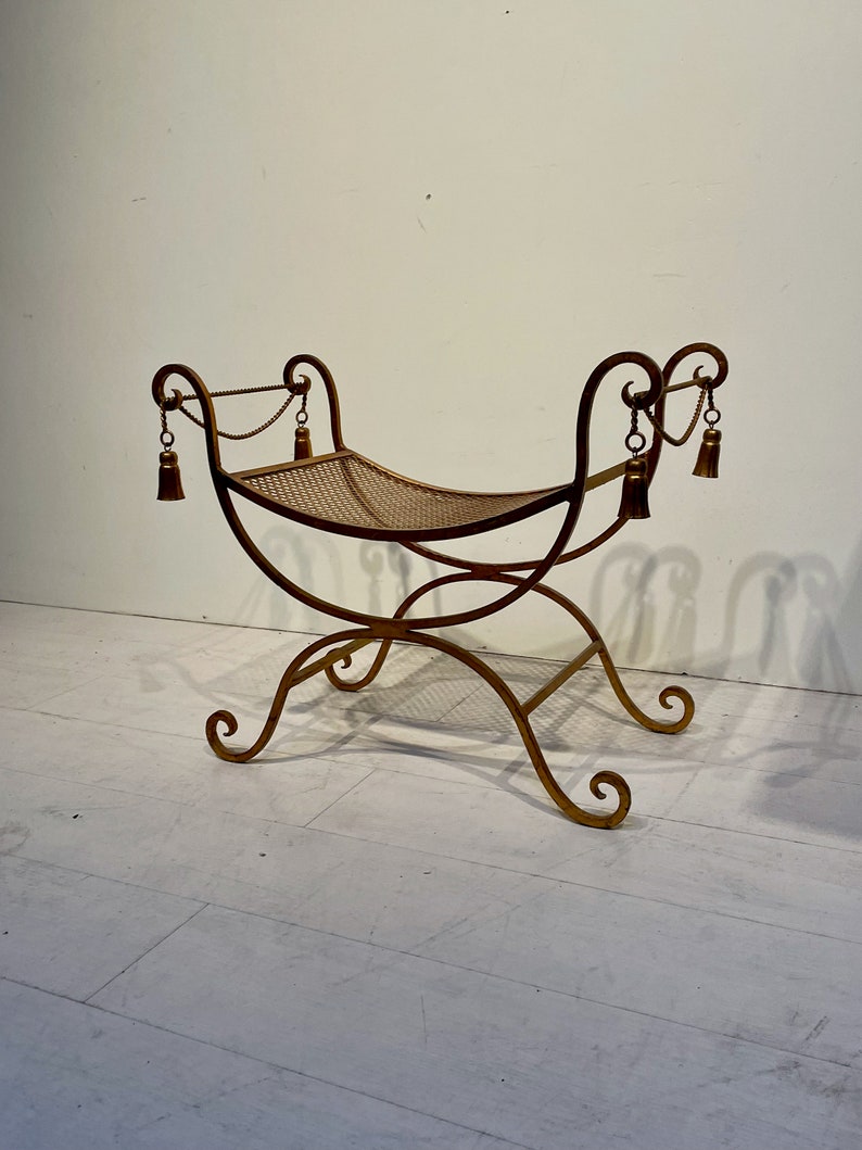 Puede incluir: Un banco de metal dorado con asiento tejido. El banco tiene patas curvas y un respaldo decorativo con borlas colgando de cadenas. El dise&ntilde;o es ornamentado y elegante, adecuado para la decoraci&oacute;n de interiores.