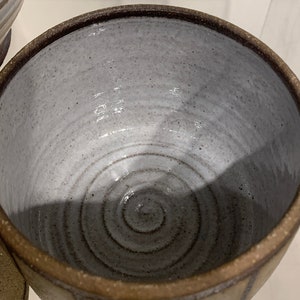 Puede incluir: Un cuenco de cer&aacute;mica hecho a mano con un interior gris claro y un borde marr&oacute;n oscuro. El exterior es de color beige claro con rayas marrones verticales. El interior del cuenco presenta un dise&ntilde;o en espiral.