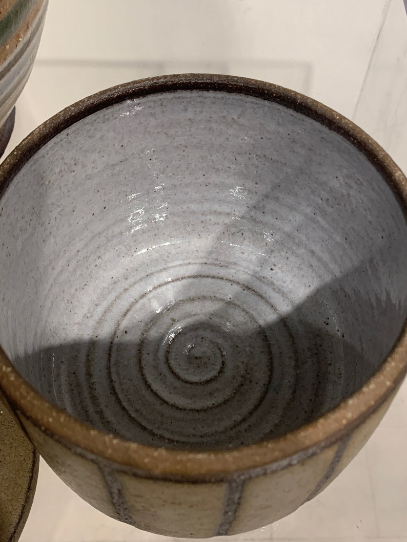 Puede incluir: Un cuenco de cer&aacute;mica hecho a mano con un interior gris claro y un borde marr&oacute;n oscuro. El exterior es de color beige claro con rayas marrones verticales. El interior del cuenco presenta un dise&ntilde;o en espiral.