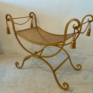 Puede incluir: Un banco de metal dorado con asiento y patas curvadas. El banco tiene borlas decorativas colgando de la parte superior y un asiento de malla. El banco est&aacute; dise&ntilde;ado con una base y reposabrazos enrollados.