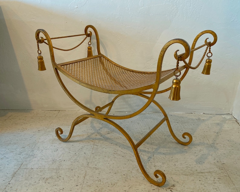 Puede incluir: Un banco de metal dorado con asiento y patas curvadas. El banco tiene borlas decorativas colgando de la parte superior y un asiento de malla. El banco est&aacute; dise&ntilde;ado con una base y reposabrazos enrollados.