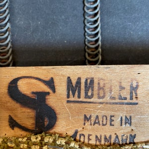 May include: Close-up of a wooden furniture component with the text "M&Oslash;BLER MADE IN DENMARK". The wood has a dark "S" logo and is trimmed with a woven fabric in shades of green, brown, and beige. The background shows a dark fabric and metal springs.