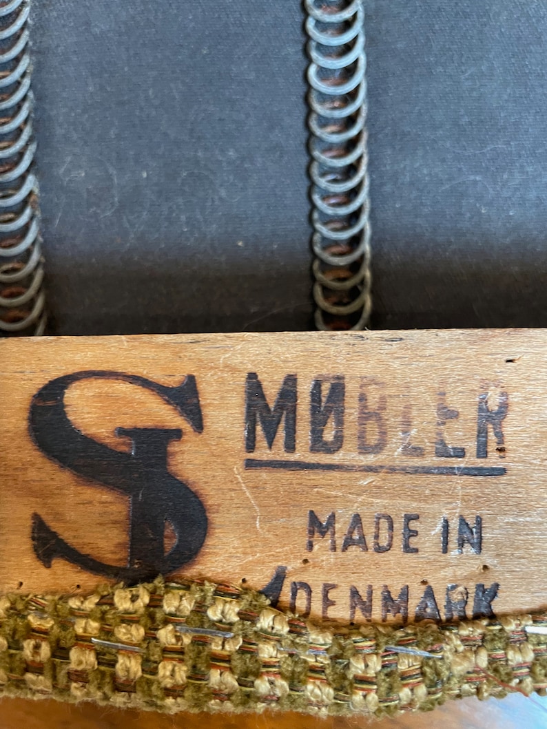 May include: Close-up of a wooden furniture component with the text "M&Oslash;BLER MADE IN DENMARK". The wood has a dark "S" logo and is trimmed with a woven fabric in shades of green, brown, and beige. The background shows a dark fabric and metal springs.