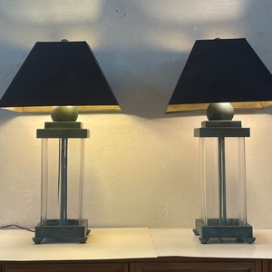 May include: Two matching table lamps with black lampshades and green bases. The bases are made of metal and have a clear glass cylinder in the center.