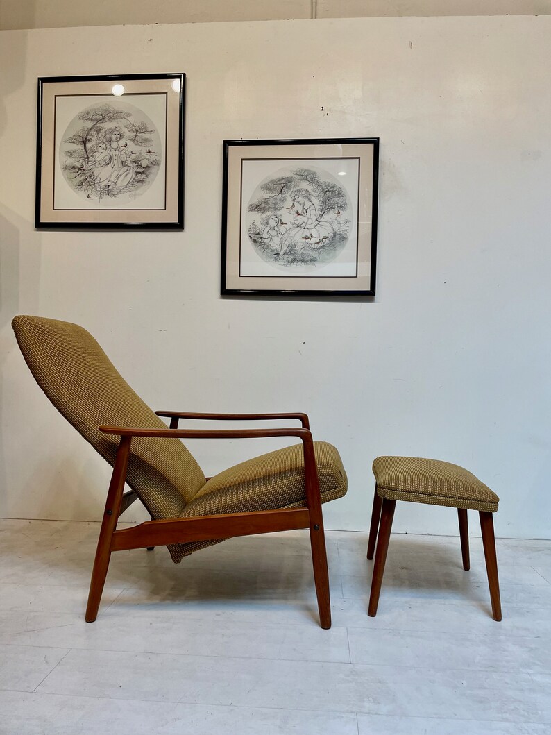 May include: A mid-century modern lounge chair and ottoman set. The chair has a high back and angled arms, with a brown fabric seat and back. The frame is made of wood. The ottoman has a matching fabric top and wooden legs. Two framed circular prints hang on the wall.
