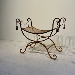 Puede incluir: Un banco de metal dorado con asiento tejido. El banco tiene patas curvas y adornos decorativos. Peque&ntilde;as borlas cuelgan de la parte superior del banco. El banco mide aproximadamente 76 cm de largo.
