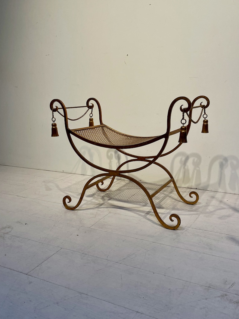 Puede incluir: Un banco de metal dorado con asiento tejido. El banco tiene patas curvas y adornos decorativos. Peque&ntilde;as borlas cuelgan de la parte superior del banco. El banco mide aproximadamente 76 cm de largo.