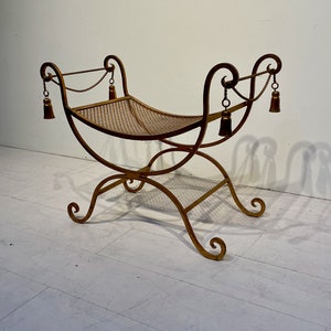 Puede incluir: Un banco de metal dorado con un asiento tejido. El banco tiene patas curvas y adornos decorativos en los brazos. Borlas cuelgan de los brazos. El banco mide aproximadamente 91 cm de largo.