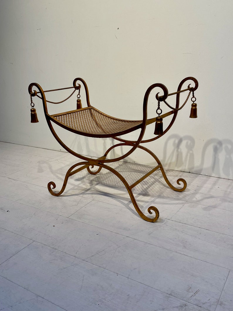 Puede incluir: Un banco de metal dorado con un asiento tejido. El banco tiene patas curvas y adornos decorativos en los brazos. Borlas cuelgan de los brazos. El banco mide aproximadamente 91 cm de largo.