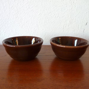 May include: Two brown ceramic bowls with a glossy finish. The bowls have a simple, rounded design and are set on a wooden surface. The bowls are empty and ready to be filled with food.