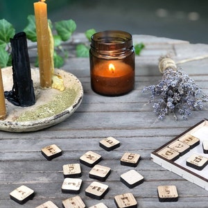 May include: Wooden tiles with Hebrew letters and numbers on them. The tiles are arranged on a wooden surface with a candle and a jar of candles in the background.