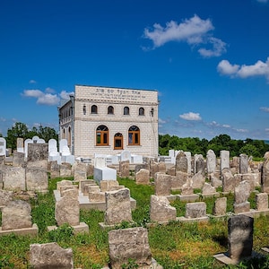 Puede incluir: Un edificio blanco con una inscripción en hebreo sobre la entrada se encuentra en medio de un cementerio judío. El cementerio está lleno de lápidas, muchas de las cuales están hechas de piedra gris.