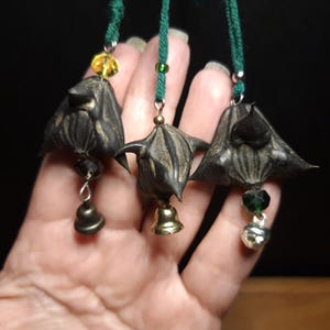 May include: Three brown, dried, water chestnut pods, each strung on a cord with a bell and a bead. The cords are green and the beads are green, yellow, and clear.