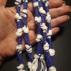 May include: Close-up of several strands of handmade macrame cord. The cords are a combination of white and blue, with knots tied at intervals. The cords are held in a hand, and the background is dark.