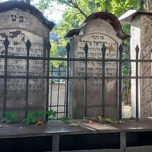 Puede incluir: Una valla de metal negro rodea una fila de tres lápidas de piedra gris con escritura hebrea. Las lápidas están en un cementerio.