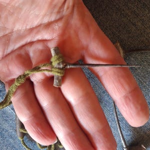 Puede incluir: Una herramienta hecha a mano con un mango de madera en forma de cruz envuelto con hilo verde. Una aguja metálica larga y delgada se extiende desde el mango. La herramienta se sostiene en una mano, con un fondo de mezclilla.