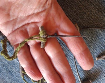 May include: A handmade tool featuring a wooden cross-shaped handle wrapped with green twine. A long, thin metal needle extends from the handle. The tool is held in a hand, with a denim background.