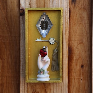 May include: A rectangular gold-painted wooden shadow box containing a silver-toned metal keyhole plate, a silver skeleton key with a beige tassel, and a white ceramic hand sculpture holding a small red glass heart ornament.