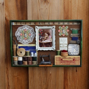 May include: A green wooden shadow box with a variety of vintage sewing and needlework items, including spools of thread, thimbles, a sewing machine, and a lace doily. The box also features a framed image of a woman sewing, and a variety of vintage labels and advertisements.