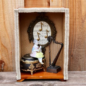 May include: A wooden shadow box with a gold-framed oval mirror with musical notes inside. A porcelain figurine of a woman playing a lute sits on a black music box with gold accents. A miniature harp sits on the right side of the box.