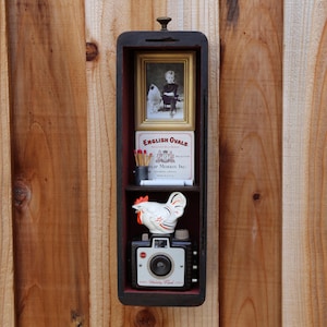May include: A black, rectangular shadow box with a small handle at the top. Inside are three shelves holding a framed photo, a box of "English Ovals" cigarettes, a ceramic rooster, and a vintage camera.