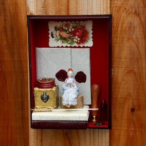 May include: A small wooden box with a red velvet interior. Inside the box is a porcelain doll, a book, a quill pen, an inkwell, a wax seal, and a card with a red rose and a hand holding a bouquet of flowers. The card reads "With love and best wishes".