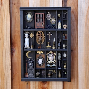 May include: A black shadow box filled with miniature gothic and antique items. The collection includes a doll, a skull, a key, a cross, a moon, a bat, a framed face, and other curiosities. The items are arranged in individual compartments.