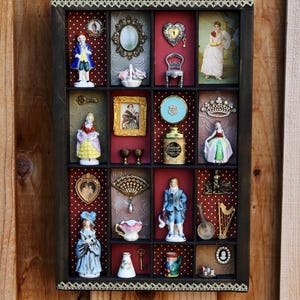 May include: A dark wooden shadow box display case with sixteen compartments containing various miniature porcelain figurines, vintage-style framed portraits, small metal decorative items, a tiny chair, a fan, a harp, and a small lidded container.