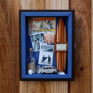 May include: A shadow box with a blue interior, featuring a wooden surfboard, vintage postcards, a seashell, a bottle with sand, and a miniature van. The box has the text "Welcome To Surf City".
