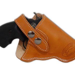 Op de afbeelding: Een bruine leren holster met een zwart revolver erin. De holster heeft een drukknopsluiting en is gestikt met wit garen. De holster is gestempeld met "Barrow Leather Hand Crafted" en "Beaumont, OR".