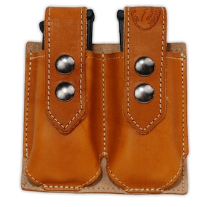 Może przedstawiać: Podwójna kabura na magazynki wykonana ze skóry brązowej z pikowanym wzorem i srebrnymi nitami. Kabura ma dwie komory na magazynki i pasek skórzany z napisem "Leather Specialties Co."