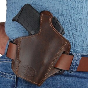 K&ouml;nnte beinhalten: Ein braunes Lederholster mit einem gen&auml;hten Design und einer G&uuml;rtelschlaufe. Das Holster ist f&uuml;r eine Pistole ausgelegt und tr&auml;gt unten das Logo "The Outfitter".