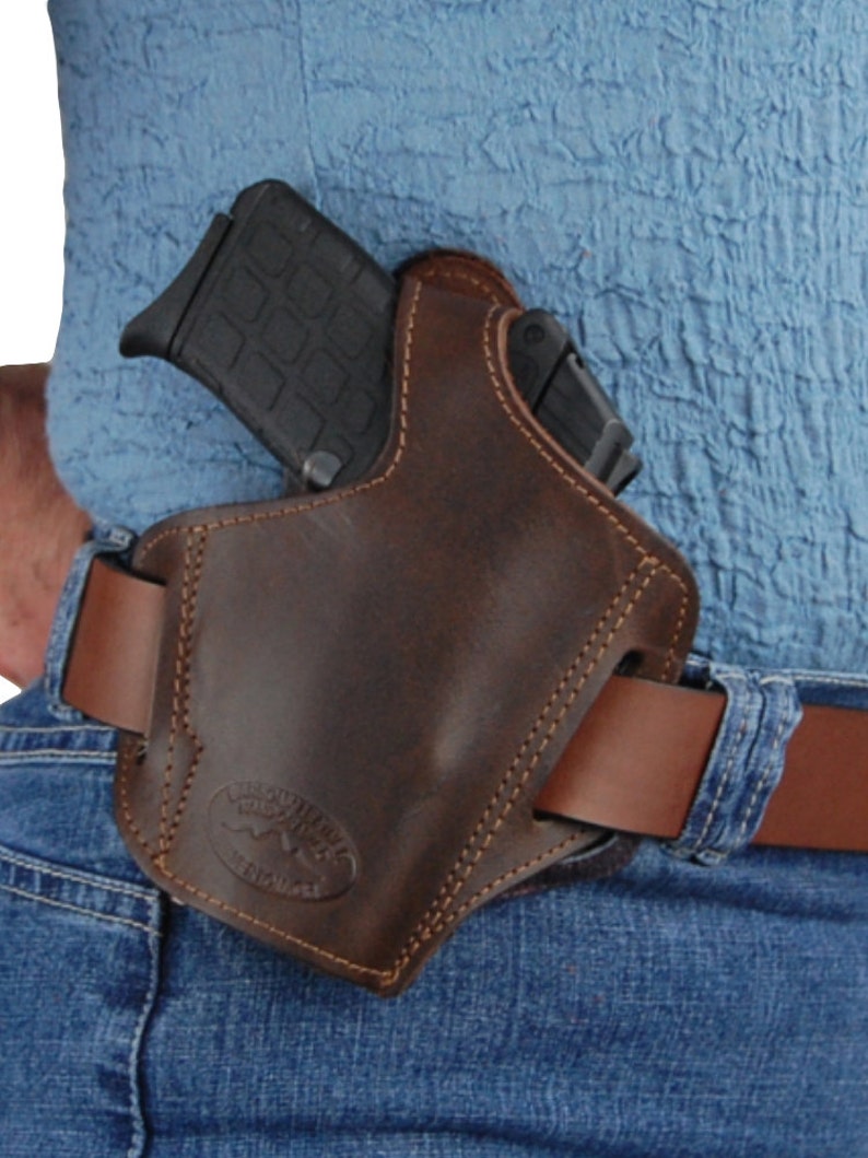 K&ouml;nnte beinhalten: Ein braunes Lederholster mit einem gen&auml;hten Design und einer G&uuml;rtelschlaufe. Das Holster ist f&uuml;r eine Pistole ausgelegt und tr&auml;gt unten das Logo "The Outfitter".
