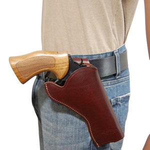 Puede incluir: Una funda de cuero marrón con costuras blancas, que sostiene un revólver con empuñadura de madera. La funda está sujeta a un cinturón negro que se lleva con vaqueros azules. La empuñadura del revólver es de color marrón claro.